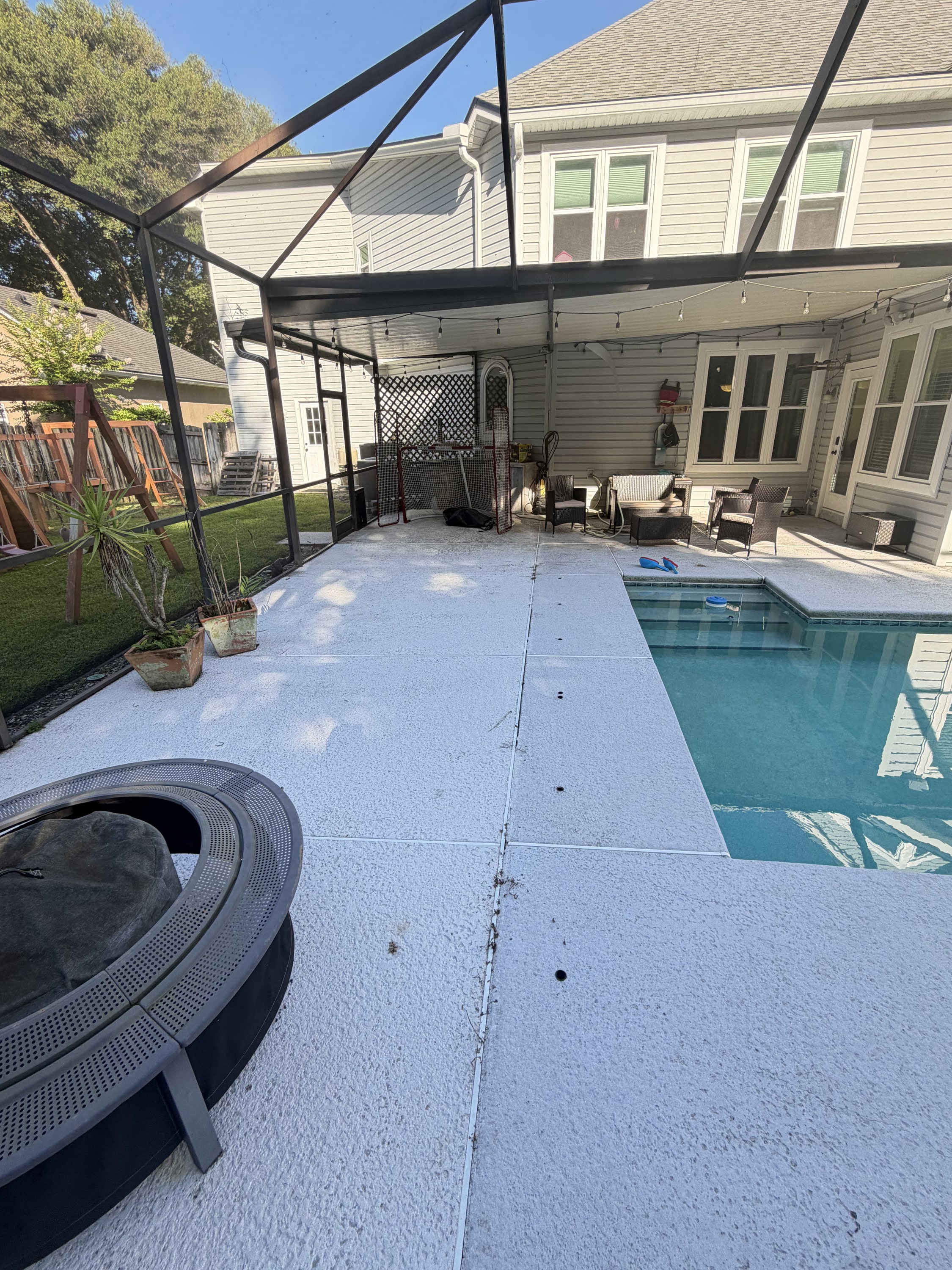 Clean light-coloured pool deck with in-ground pool, black pergola, and house.