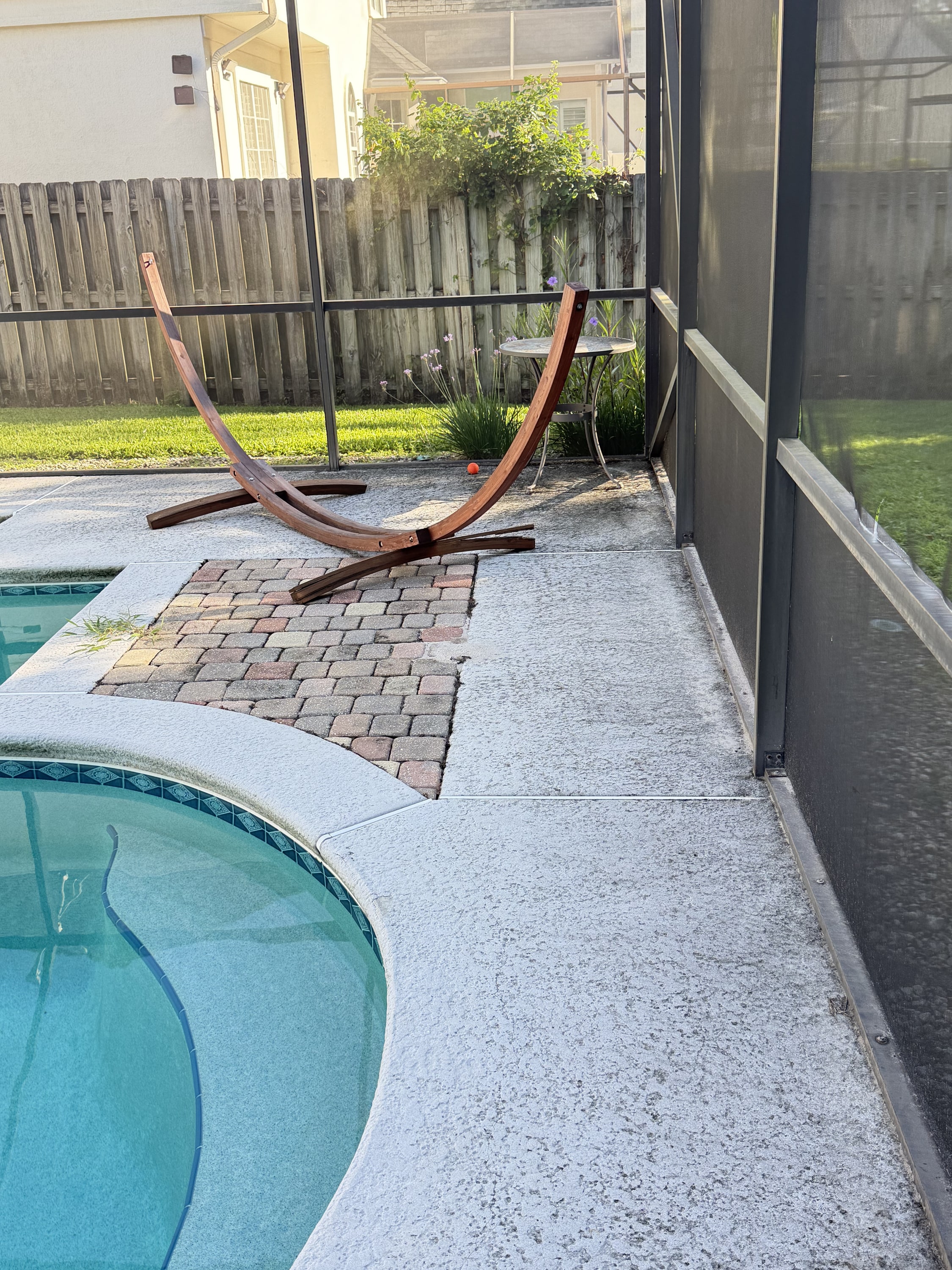 Poolside patio with new concrete surround and decorative paver section, pool and wooden fence.
