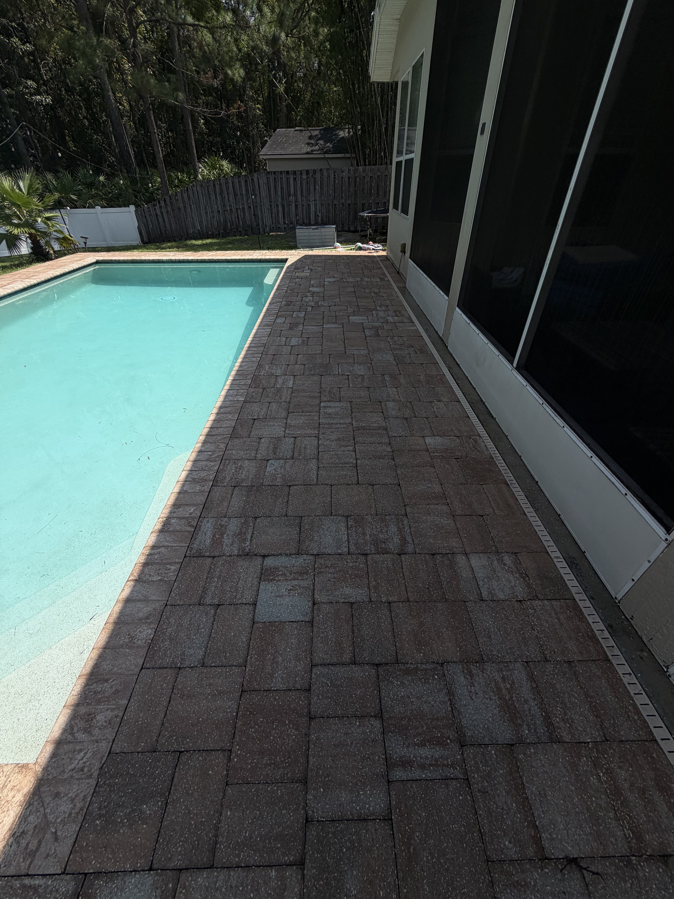 Dark block paving pathway beside a turquoise swimming pool and white house.