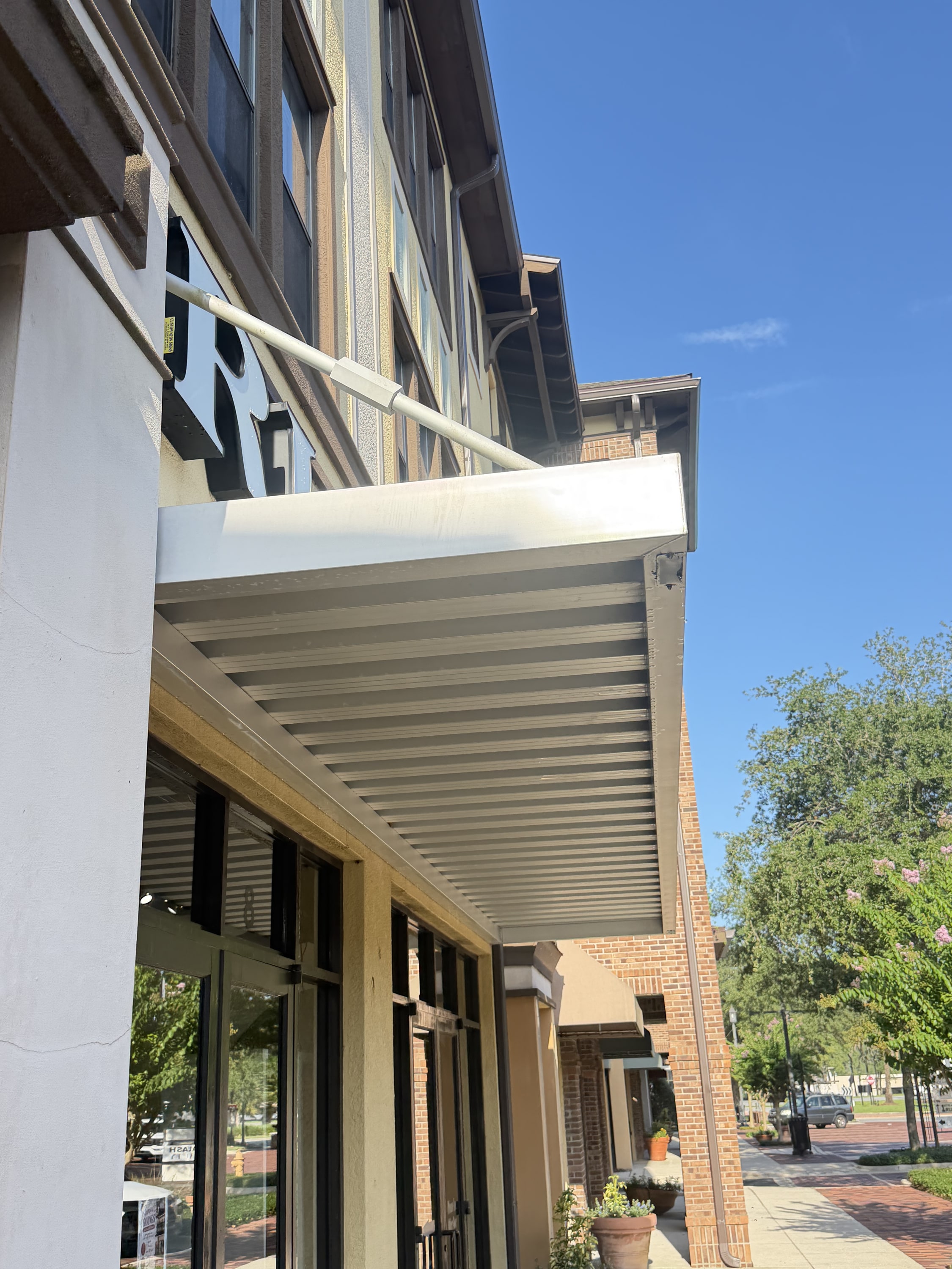 Modern white louvered pergola installed over a commercial storefront with glass doors and brick building backdrop.
