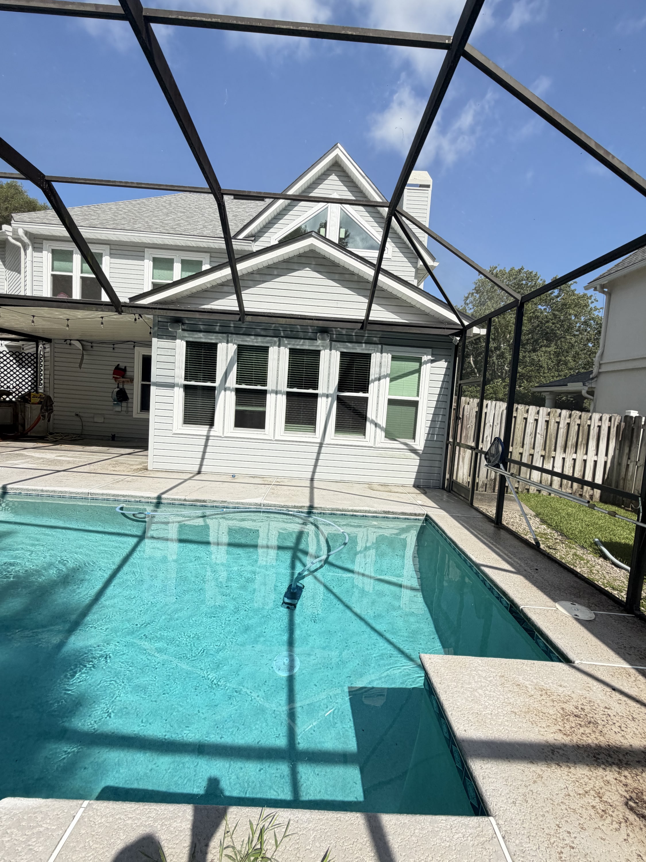 Black aluminum pool screen enclosure over turquoise pool with concrete deck and white house in background.