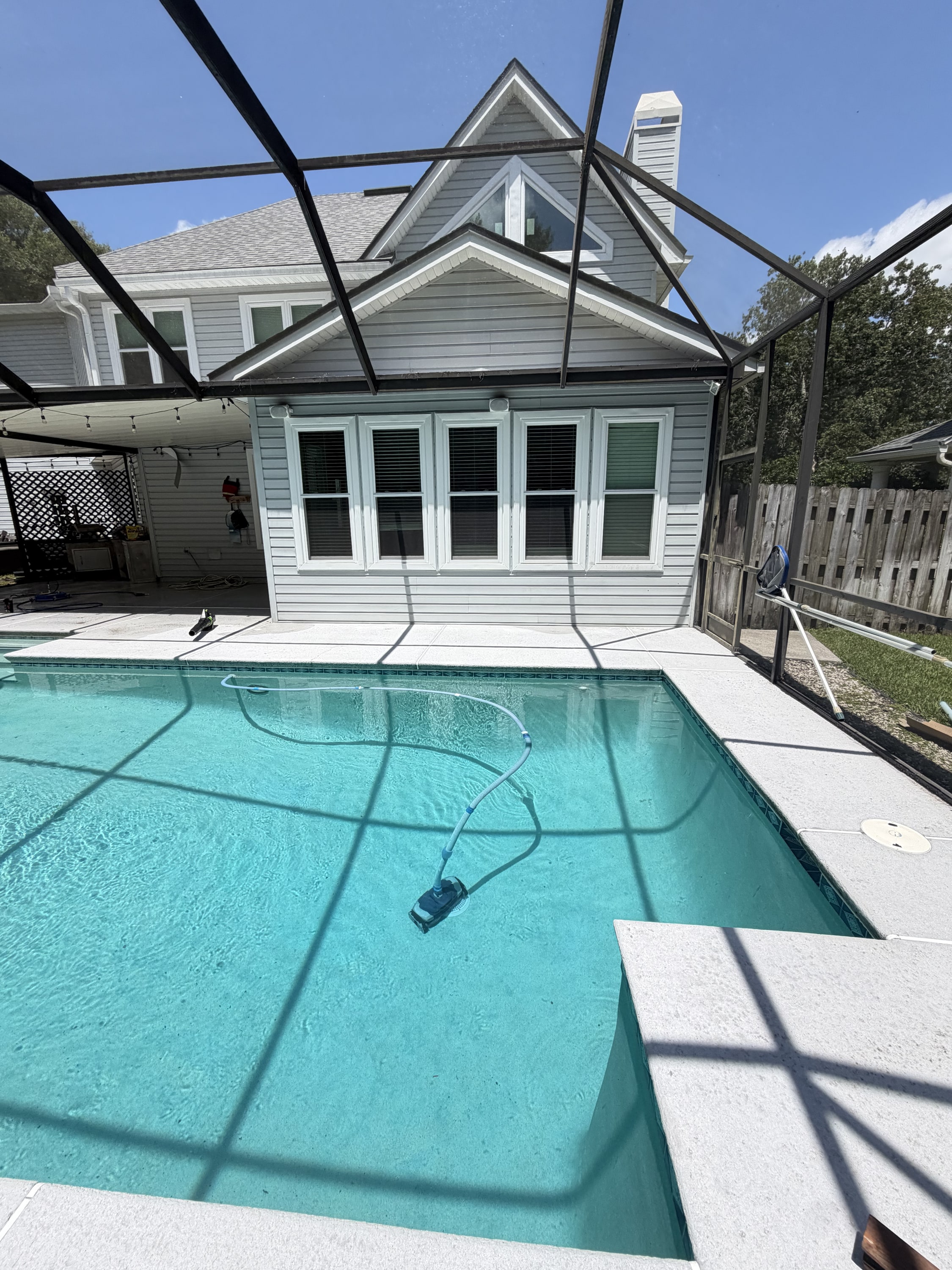 Swimming pool with black screening cage, white pool house, and concrete deck in sunny backyard.
