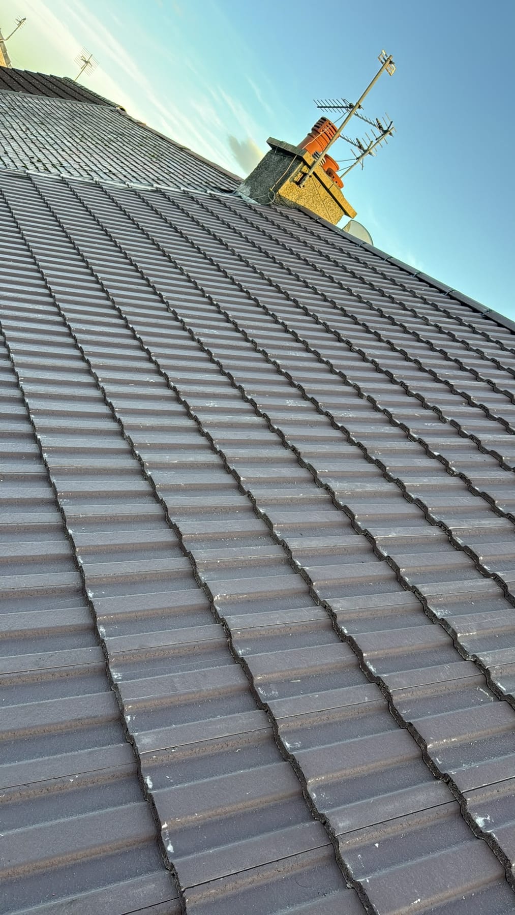 Grey interlocking roof tiles with yellow chimney and TV aerials shot from below against blue sky.